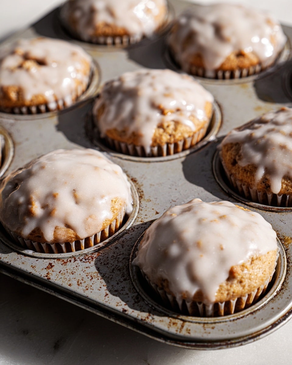 The image shows a metal muffin tray with eight baked muffins, each topped with a light, shiny white icing glaze that slightly drips down the sides. The muffins have a textured, somewhat rough surface with small, round pieces visible on top, indicating a crumbly or nutty texture beneath the smooth icing. The muffins fill each cup of the tray closely, creating rounded tops that rise a little above the edges. The tray itself is slightly worn with some marks, placed on a white marbled surface with soft natural light creating gentle shadows on the left side. photo taken with an iphone --ar 4:5 --v 7