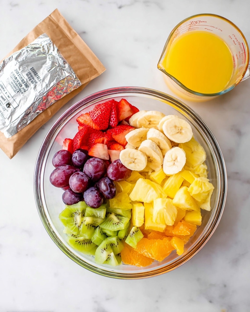 A clear bowl holds a colorful mix of six fruit layers arranged side by side: red sliced strawberries, purple grapes (some halved), pale yellow banana slices in the center, yellow pineapple chunks, bright orange pieces, and green sliced kiwi at the bottom left. Below the bowl on a white marbled surface, there is a silver foil package, a brown paper bag, and a glass measuring cup filled with orange juice. The overall scene is bright and fresh with the bowl centered on the surface. photo taken with an iphone --ar 4:5 --v 7